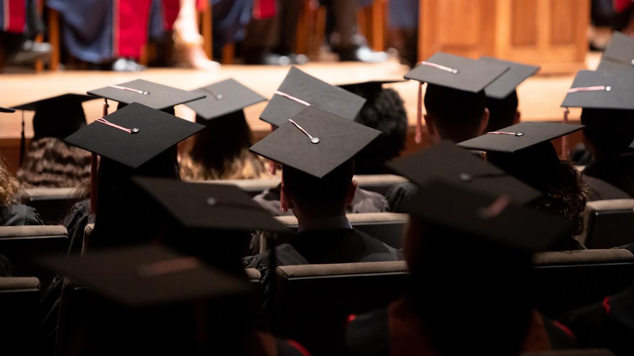 毕业典礼上的一群毕业生. 他们戴着传统的黑色帽子，穿着长袍. 背景是爱丽丝塔利大厅，一排排的座位上坐满了毕业生. 这张照片的角度说明是从大厅后面拍的, 重点是毕业生的后脑勺和学位帽, 在前景中有一个作为焦点.